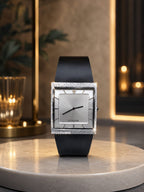 A rectangular STOLIDE watch with a black leather strap, a silver dial, and a diamond-encrusted bezel, displayed on a round marble pedestal in a warm, upscale interior setting.