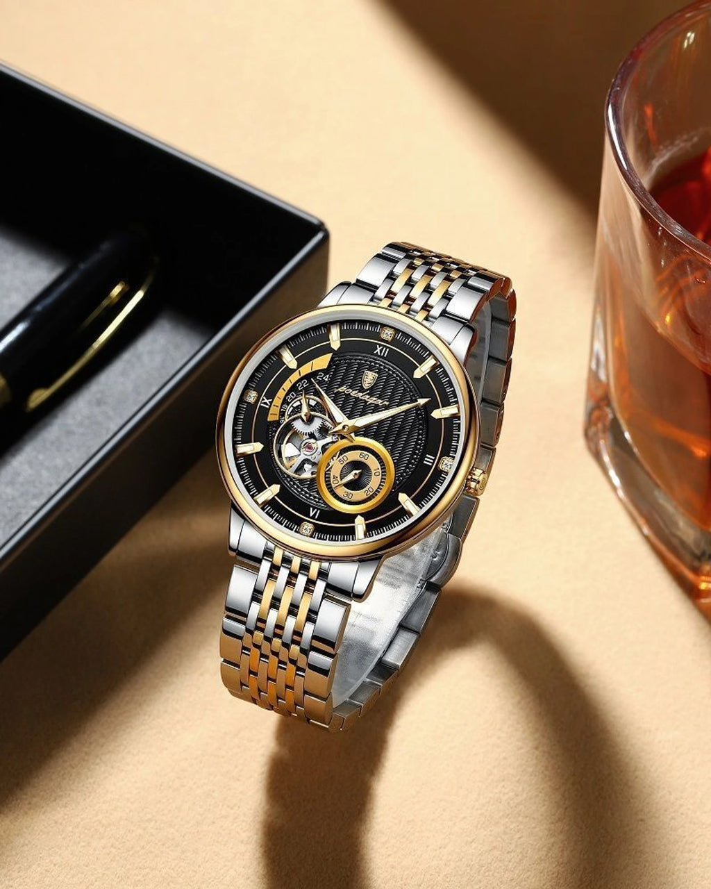 A two-tone stainless steel and gold automatic watch with a black skeleton dial, displayed on a beige surface next to a black box and a glass of amber liquid.