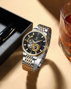 A two-tone stainless steel and gold automatic watch with a black skeleton dial, displayed on a beige surface next to a black box and a glass of amber liquid.