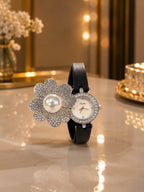 A luxury wristwatch with a black leather strap, featuring a silver-tone flower-shaped bracelet embellished with crystals and a central white pearl, alongside a round white dial watch face encrusted with crystals. The watch is displayed on a polished marble tabletop with warm bokeh background lights.