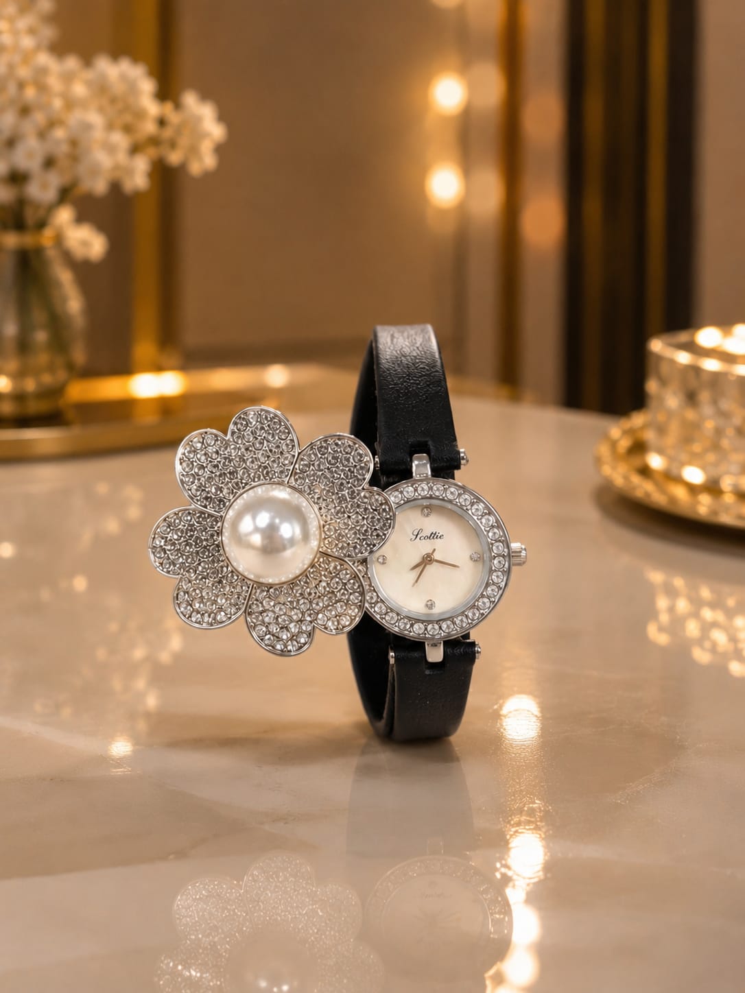 A luxury wristwatch with a black leather strap, featuring a silver-tone flower-shaped bracelet embellished with crystals and a central white pearl, alongside a round white dial watch face encrusted with crystals. The watch is displayed on a polished marble tabletop with warm bokeh background lights.