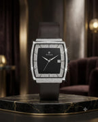 A luxury men's watch by TITAN, featuring a square black dial, a bezel encrusted with sparkling diamonds, and a dark brown leather strap. The watch is displayed on a polished black marble pedestal in an elegant, dimly lit room with warm ambient lighting.