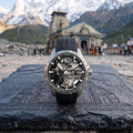 A luxury skeleton wristwatch with a black strap and a silver-toned case, displayed on a dark stone slab with a blurred mountain temple background.