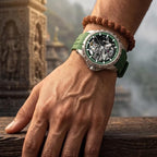 A close-up of a man's hand resting on a wooden surface, wearing a green rubber strap watch with a detailed skeleton dial and a brown beaded bracelet.