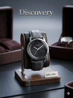 A Discovery wristwatch with a dark dial and black leather strap displayed on a wooden stand. The watch is centered against a dark gray background with blurred brown presentation boxes in the background.