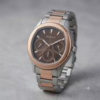 A high-resolution professional studio photograph of a two-tone men's chronograph watch from Discovery, presented against a neutral grey textured background. The watch features a silver stainless steel case and bracelet, accented by a prominent rose gold-tone bezel. The brown dial is textured with a subtle horizontal stripe pattern and displays three rose gold-rimmed sub-dials for chronograph functions. The 'Discovery' logo is positioned below the 12 o'clock marker, and the applied hour markers and hands are