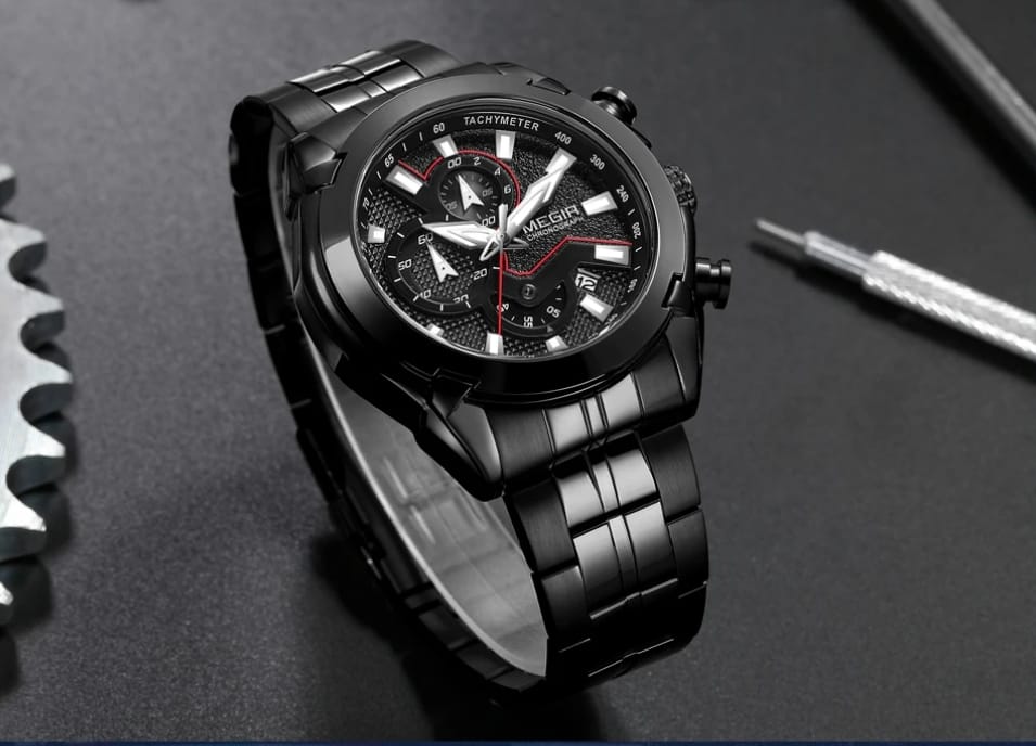 A black stainless steel wristwatch with a black dial, silver-tone hands and markers, and a thin red seconds hand. The watch is displayed on a dark textured surface with a gear on the left and a screwdriver on the right.