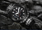 A black stainless steel chronograph wristwatch with a matching metal link bracelet, resting on dark rocks. The watch features a textured black dial with silver-tone indices and a red second hand.