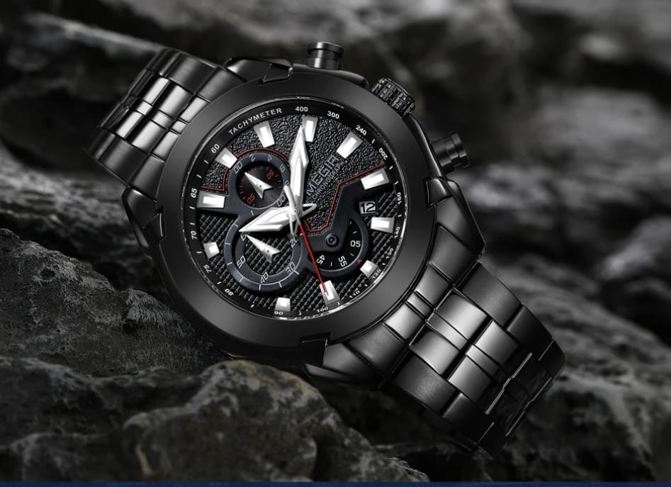 A black stainless steel chronograph wristwatch with a matching metal link bracelet, resting on dark rocks. The watch features a textured black dial with silver-tone indices and a red second hand.