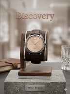 A wristwatch with a round beige dial, silver-toned case, and brown leather strap displayed on a wooden stand. The watch is set against a softly blurred indoor background with the word 'Discovery' above it and a stone pedestal below with an 'MRP ₥1599' plaque.