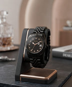 A black and rose gold wristwatch with a matching black link bracelet and textured bezel, displayed on a black and wood stand on a dark marble surface.