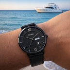 A black leather strap wristwatch with a textured black dial and silver-tone hands and markers, shown worn on a person's wrist with a blurred ocean and yacht background.