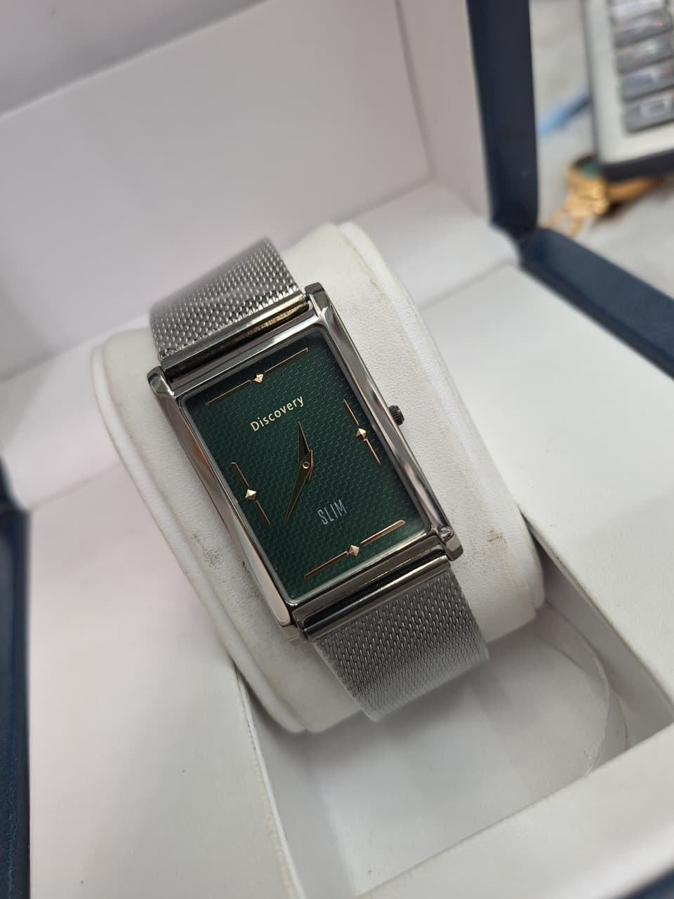 A silver-tone rectangular analog watch with a textured dark green dial and a matching silver mesh strap, displayed inside a white watch box.