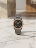 A men's analog wristwatch with a black dial, rose gold-toned bezel and hands, and a dark gray metal link bracelet, displayed on a white marble surface.