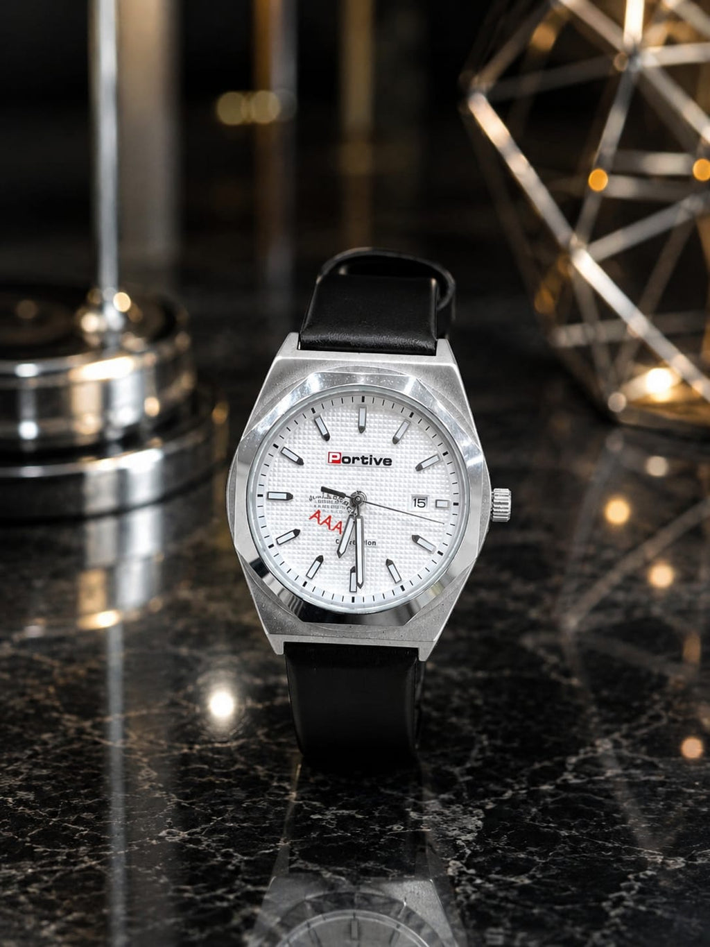 A wristwatch with a silver-toned metal case, white dial, and black leather strap, displayed on a dark marble surface with blurred decorative objects in the background.