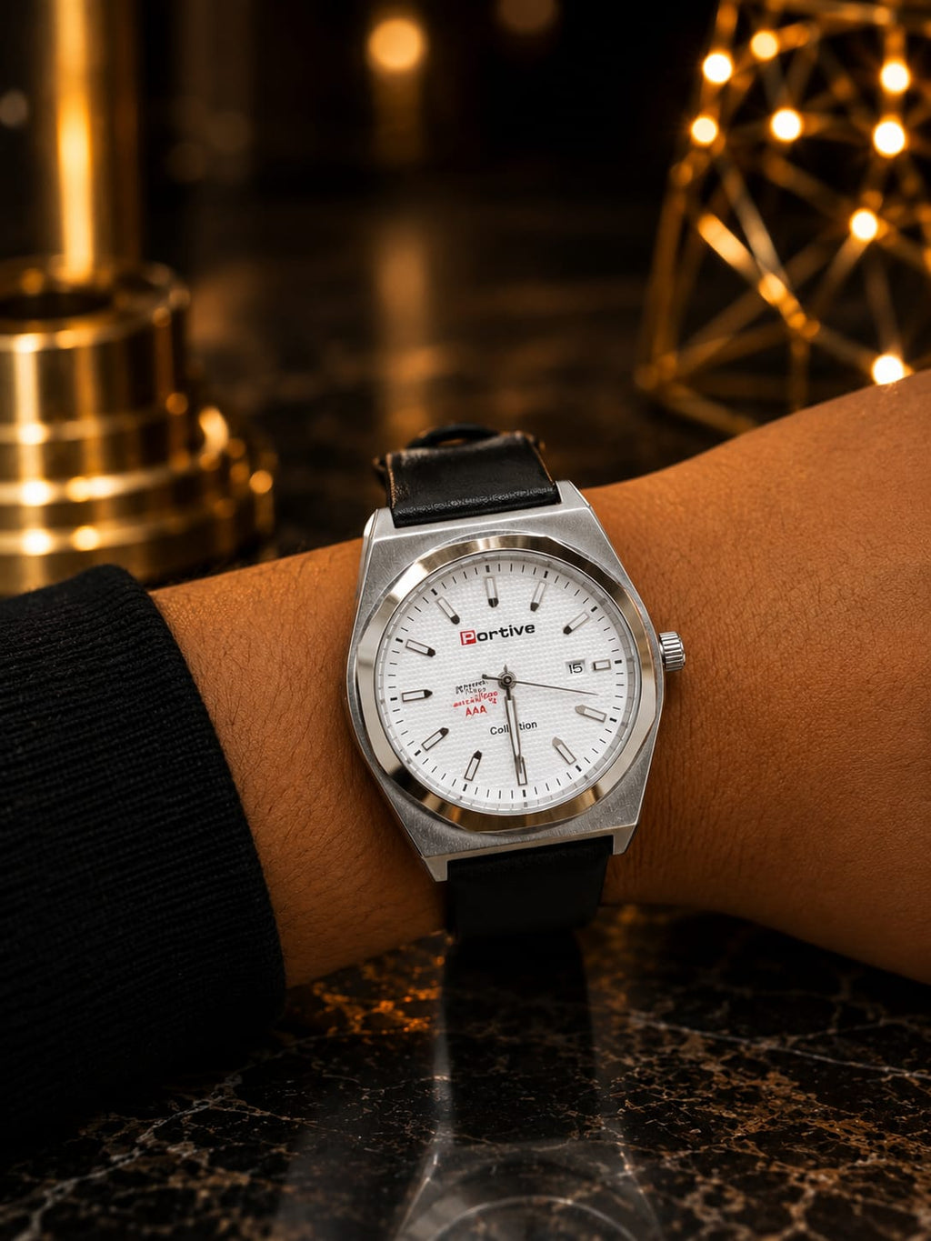 A wristwatch with a silver-toned metal case, white dial, and black leather strap worn on a person's wrist, set against a dark marble surface with warm golden bokeh lights in the background.