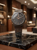 A luxury wristwatch with a dark gray dial, rose gold-tone indices and hands, and a dark gray metal case and bracelet, displayed on a clear stand atop a black marble slab in a high-end boutique setting.