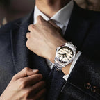 A silver-toned metal wristwatch with a beige dial and date window, worn on a man's wrist as he adjusts his tie with a dark textured blazer.