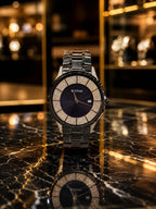 A luxury men's watch with a black dial, a bezel encrusted with sparkling clear stones, and a matching black metal link bracelet, displayed on a polished black marble surface with a warm, blurred background.